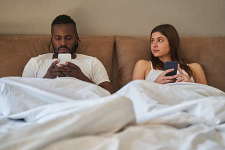 Young mixed-race couple with cellphones in bedroomの写真素材