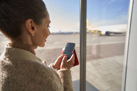 Female clicking her smartphone near airport windowの写真素材
