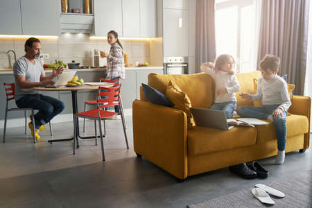 Children on sofa doing homework while parents in kitchenの写真素材