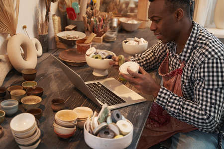 Joyous ceramist seated at laptop showing earthenware products to customer