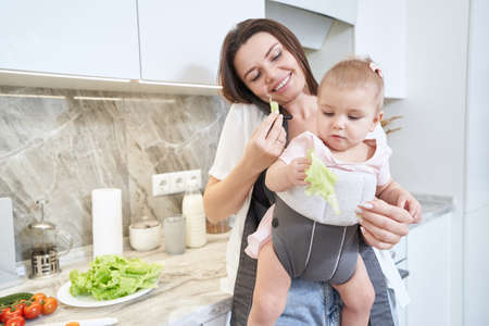 Elegant brunette mother with kid in baby carrier holding salad in hand