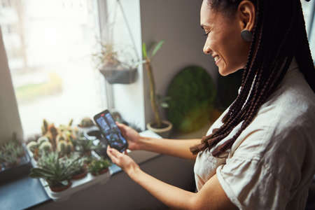 Joyous floral designer photographing potted exotic plants with her cellphone