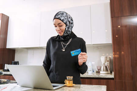 Happy muslim woman using bank card and notebook at home