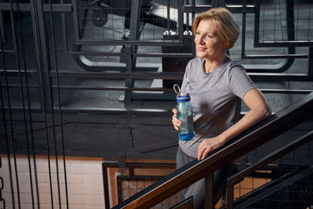 Cheerful woman with drink standing on stairs in gymの写真素材
