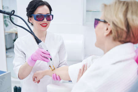 Cheerful cosmetologist removing hair from woman arm with laser device
