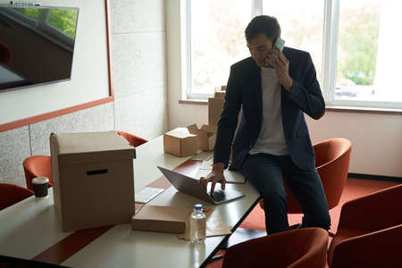 Man among unwrapped boxes using laptop during phone talkの写真素材