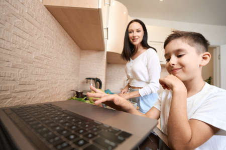 Satisfied child using laptop touchpad near cooking mother