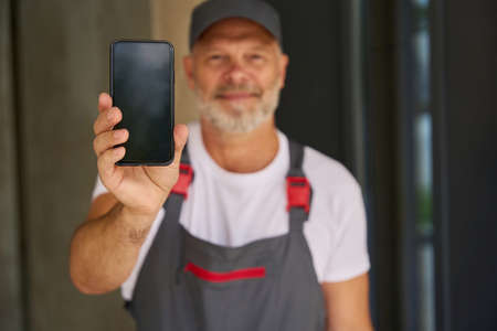 Smiling builder wearing cap and overalls shows mobile phoneの写真素材