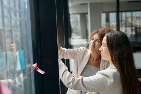 Smiling young females stand near window and tear off stickersの写真素材