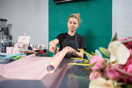 Focused florist cuts piece of pink paper in flower shop