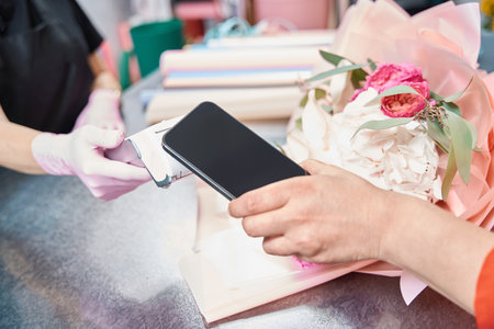 Woman holds phone near terminal to pay for ordered bouquet in flower boutique