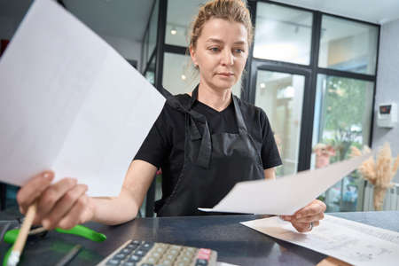 Focused florist entrepreneur is sitting at counter and checking documentsの写真素材