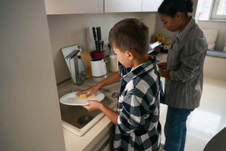 A male child is washing the dishes near the mother at homeの写真素材