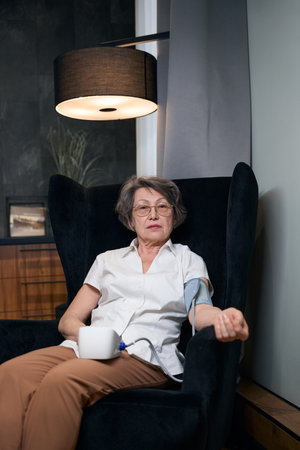 Calm old woman sits in armchair and measures her blood pressure