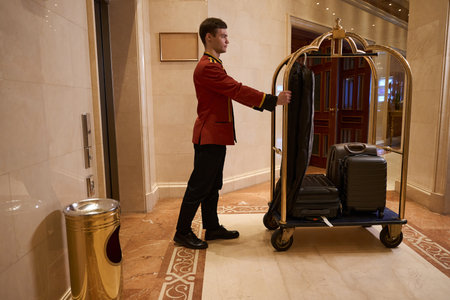 Young hotel employee is carrying cart with luggage along lobbyの写真素材