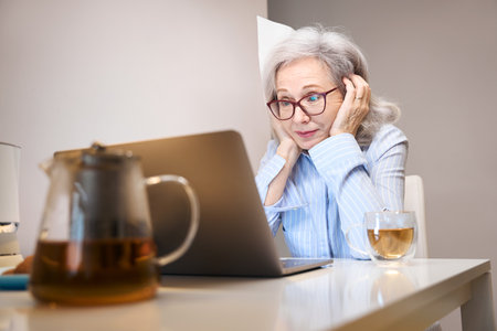 Joyful pensioner sits on a laptop at the kitchen tableの写真素材