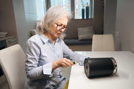 Advanced elderly female in glasses is repairing electric kettle with screwdriverの写真素材