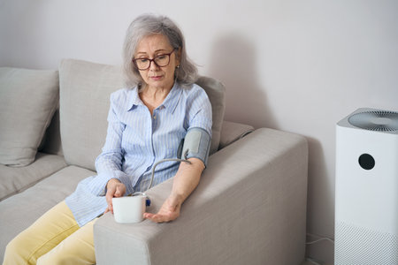 Calm elderly woman sits on couch , measures blood pressure with tonometerの写真素材