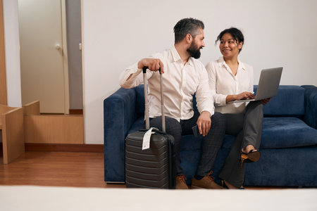 Happy mixed-race tourist couple seated on sofa in hotel roomの写真素材