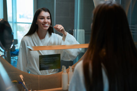 Long-haired young female brushes her teeth inbathroom in front mirror