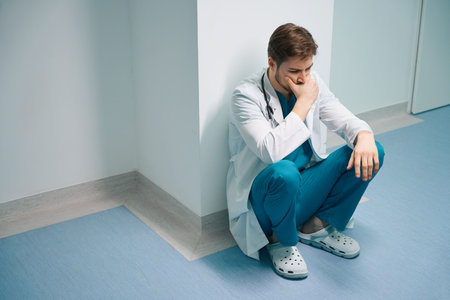Upset young surgeon squatting in hospital corridorの写真素材