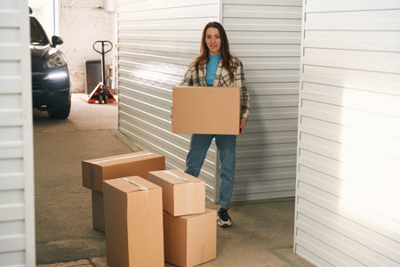 Full length of smiling woman with big cardboard boxes in self storage unit