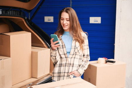 Happy lady with mobile phone and cardboard boxes in car trunk into warehouse