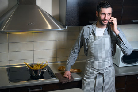 Man in chefs apron is standing in kitchen with phoneの写真素材