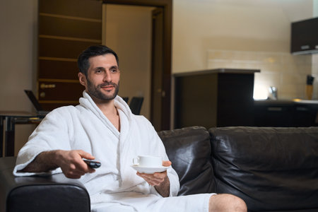Brunette with TV remote control in his hand enjoys coffeeの写真素材