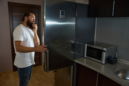 Bearded man in jeans takes something out of large refrigeratorの写真素材