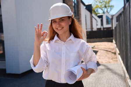 Woman architect with blueprints in hands and hardhat showing ok signの写真素材