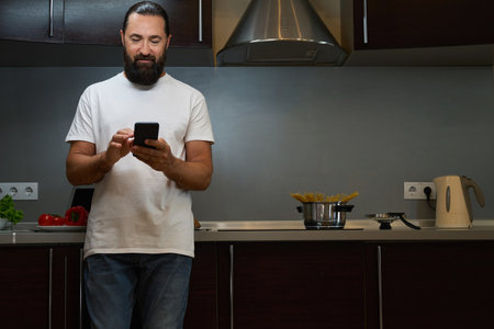 Bearded man stands in kitchen area and texts on mobile phoneの写真素材