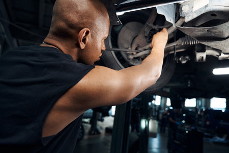 African American mechanic working under the car using long screwdriver