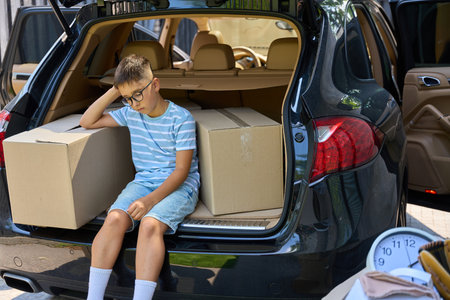 Sad boy sits in an open trunk between cardboard boxes