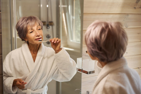 Elderly woman brushes her teeth with toothbrush in front of mirrorの写真素材
