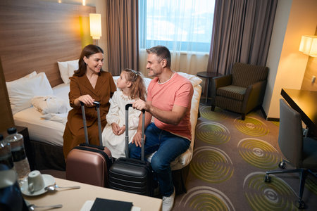Mother, father and daughter with suitcases sitting on bed in hotel roomの写真素材