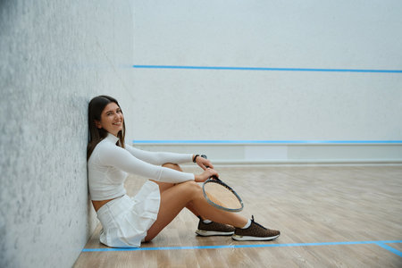 Athletic cheerful woman holding squash racket in her hands sittingの写真素材