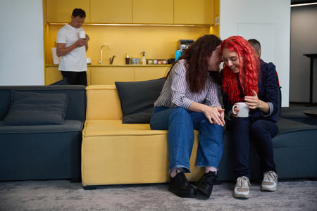 Two girls whispering and drinking coffee on soft couch in office