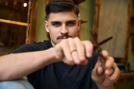 Coiffeur showing scissors while sitting at his workplace in barbershop