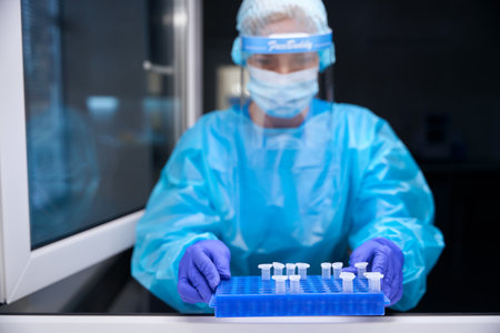 Woman in protective equipment holds organizer with test tubes in her hands