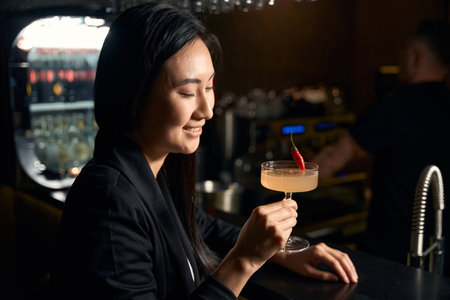 Smiling Asian woman enjoying vanilla-sweet cocktail sitting at bar counterの写真素材
