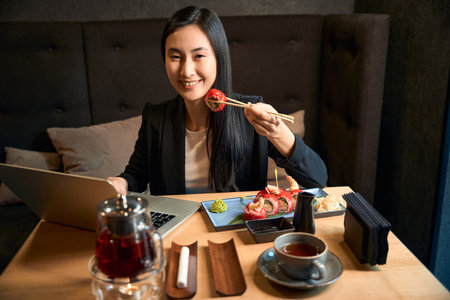 Positive asian woman holding sushi with chopsticks sitting on table with laptopの写真素材