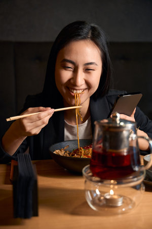 Happy Asian woman sincerely smiling eating and chatting on smartphoneの写真素材