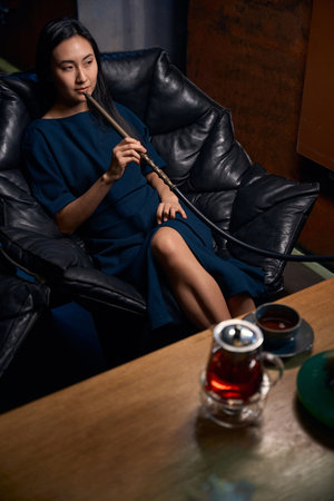 Relaxed Asian woman sitting in leather armchair smoking hookah