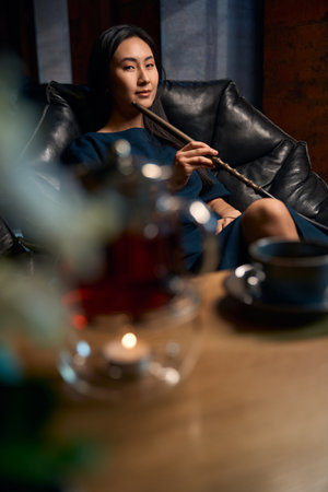 Self-confident attractive woman smoking hookah sitting in big cozy armchair