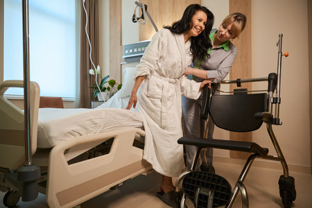 Caring nurse helps clinic patient transfer from bed to wheelchairの写真素材