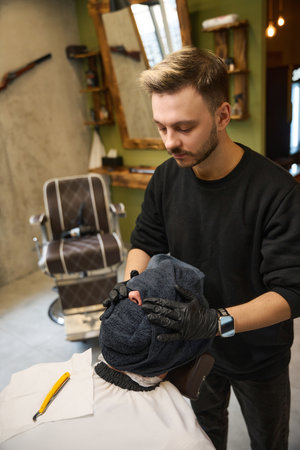 Barber tenderly massaging male face wrapped in hot towelの写真素材