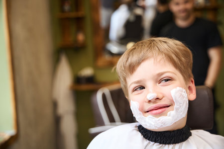 Little boy sitting in barber chair with shaving cream on faceの写真素材