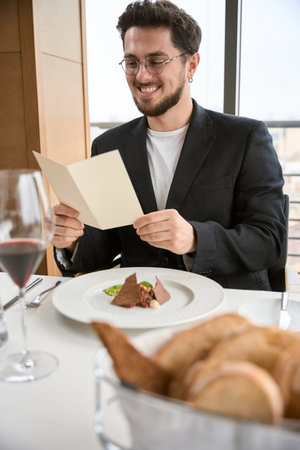 Happy young man in black suit and glasses having lunch, holding bill while sitting in the restaurantの写真素材