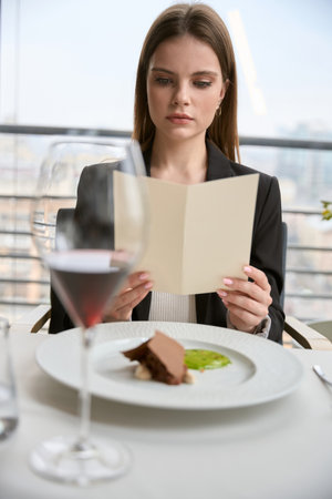 Serious young woman in black jacket having lunch, holding bill while sitting in the restaurantの写真素材